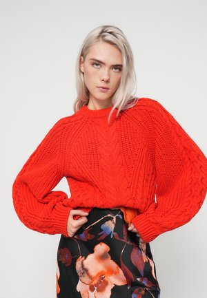 Rode cable-knit trui met brede, gestructureerde mouwen en een ronde hals, gecombineerd met een zwart rokje met bloemmotief.