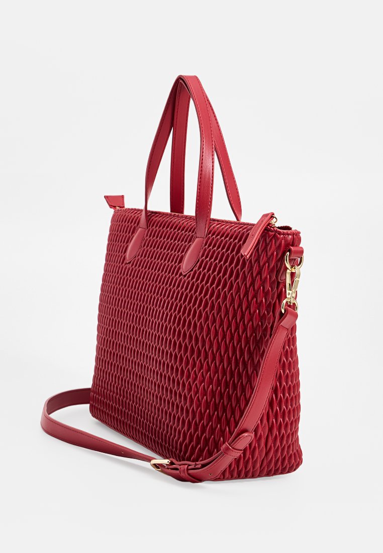 Sac à épaule texturé rouge avec deux poignées et une bandoulière réglable, quincaillerie en ton doré, se tenant contre un fond blanc.