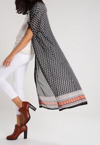 Kimono de estampado en blanco y negro con mangas largas, que presenta un borde en naranja y blanco. Se lleva con jeans blancos y botas de tacón marrones.