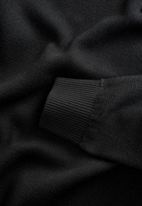 Črn pleten sweater material, ki prikazuje gladko teksturo, z detajlom rebrastega zapestja. Ima enotno barvo in mehak padec.