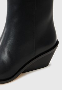 Stivale a zeppa in pelle nera che mette in evidenza una texture liscia, una forma affusolata e una leggera lucentezza. Presenta un tacco impilato con una finitura opaca.