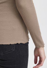 Beige, geripptes Langarmoberteil mit gewelltem Saum und Ärmelbündchen, kombiniert mit dunklen Jeans. Hat einen figurnahen Schnitt und eine weiche Textur.