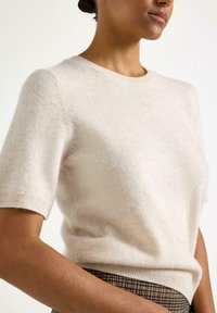 Beige kortærmet sweater lavet af blødt strikket materiale. Rund halsudskæring og tætsiddende pasform med ribkant. Stylet med bukser i ternet mønster.