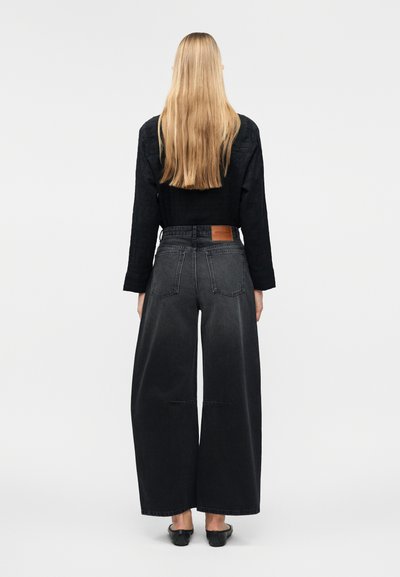 Γυναίκα με μακριά ξανθά μαλλιά που φοράει μαύρο μπλουζάκι με μακριά μανίκια, φαρδιά ξεθωριασμένα μαύρα τζιν και μαύρα παπούτσια χωρίς κορδόνια, στέκεται με την πλάτη προς τα εμπρός σε λευκό φόντο.