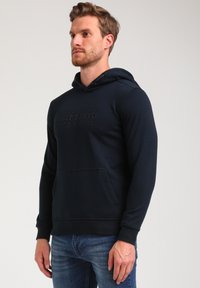 Sudadera azul marino hecha de una mezcla de algodón con un bolsillo tipo canguro, que presenta detalles de logo en relieve en el pecho y puños de canalé.
