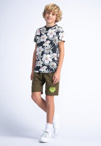 T-shirt noir à manches courtes avec des fleurs roses et blanches, associé à un short vert olive avec un logo jaune, des chaussures blanches et des chaussettes mi-mollet.