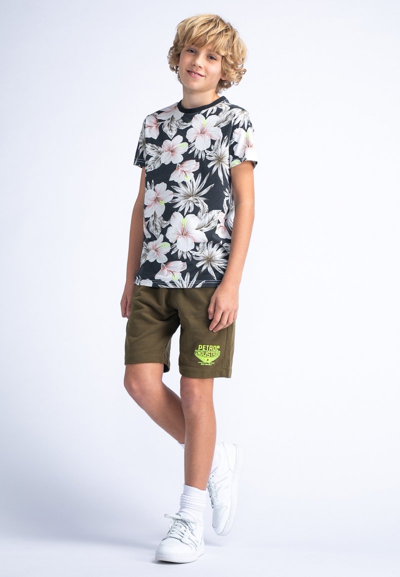 T-shirt noir à manches courtes avec des fleurs roses et blanches, associé à un short vert olive avec un logo jaune, des chaussures blanches et des chaussettes mi-mollet.