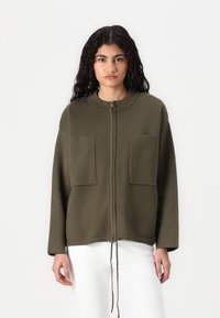 Olijfgroen ritsjack van zacht materiaal, met ribbelaccenten op de voorzakken en een trekkoord aan de onderkant voor een verstelbare pasvorm.