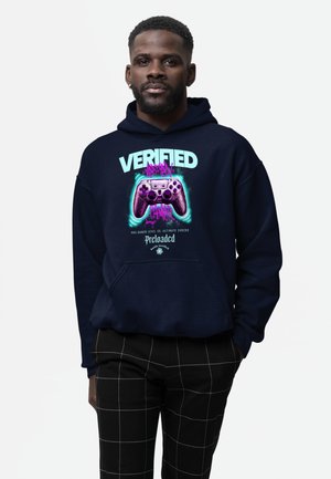Man draagt een marineblauwe hoodie met een neon afbeelding van een videogamecontroller en de tekst "Verified Virtual Gamer Reloaded," gecombineerd met zwarte geruite broek.