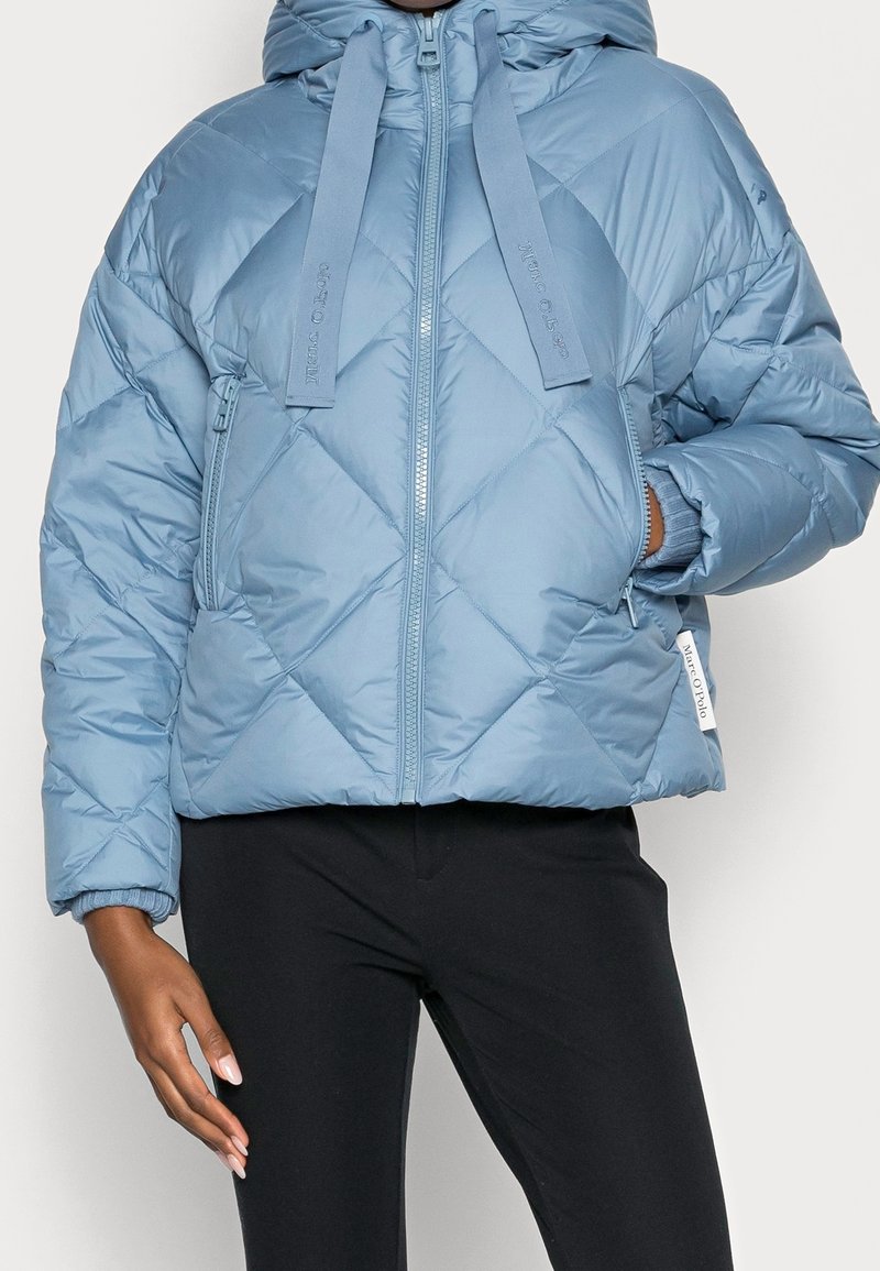 Personne portant une doudoune matelassée bleu clair avec capuche, fermeture éclair frontale, grands cordons et poches latérales, associée à un pantalon noir.