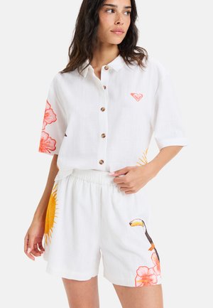 Chemise en lin blanc avec des imprimés floraux orange et un logo en forme de cœur ; short à taille élastique présentant un design de toucan et de soleil.
