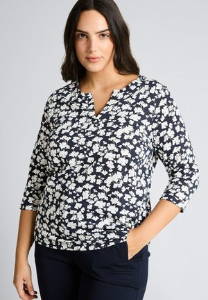 Kobieta ubrana w czarno-białą bluzkę w kwiaty z rękawami do łokcia, stojąca z jedną ręką w kieszeni na jednolitym tle.