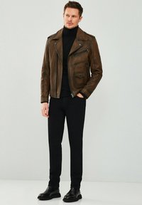 Veste de moto en cuir marron avec fermeture éclair sur le devant, poches zippées et col en pointe ; pull noir, pantalon noir et bottes noires.
