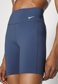 Detailní záběr na osobu, která má na sobě vysoké modré sportovní šortky s logem Nike a bílé sportovní podprsenky, stojící se uvolněnými pažemi.