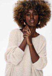 Beżowy oversize sweter o miękkiej fakturze i ściągaczach przy mankietach. Modelka ma kręcone włosy i nosi duże złote kolczyki w kształcie koła.