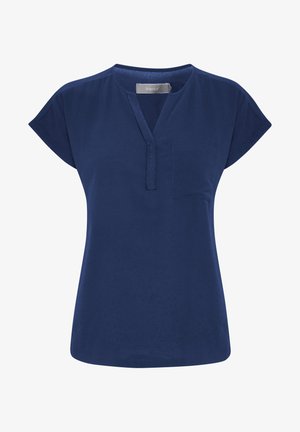 Kvinde stående iført en mørkeblå kortærmet bluse, hvide smalle bukser og tan-farvede flade sko, mod en ensartet lys baggrund.