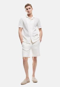 Maglietta bianca a maniche corte con texture e bottoni, abbinata a pantaloni corti bianchi. Il modello indossa scarpe beige senza lacci. Completo semplice e monocromatico.