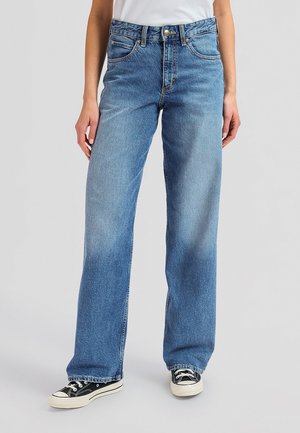 Άτομο που φοράει μπλε τζιν με ευθείες ρίγες, λευκό T-shirt και μαύρα χαμηλά αθλητικά παπούτσια, στέκεται σε γκρι φόντο.