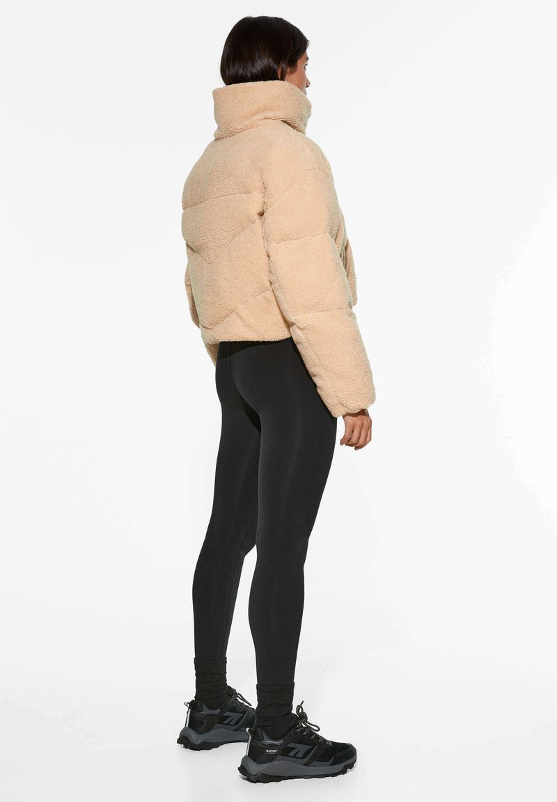 Beige kurz geschnittene Daunenjacke mit hohem Kragen, strukturiertem Stoff und Zickzackmuster. Getragen über schwarzen Leggings und schwarzen Sneakers.