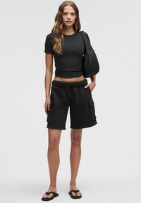 Tricou negru ajustat, pantaloni scurți cargo negri cu șnur și buzunare laterale, model purtând sandale negre de tip slide, design simplu, accente minime.