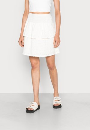 Kvinde iført hvid lagdelt mininederdel med smock-talje, hvid cropped top og hvide slid-sandaler med sorte såler.