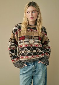 Pull en tricot avec un motif géométrique dans des tons de marron, rouge et crème. Il a un col rond et des manches amples.