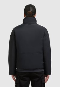 Mężczyzna w czarnej, pikowanej kurtce oraz czapce bejsbolowej z siateczki, pokazany od tyłu na jednolitym szarym tle.