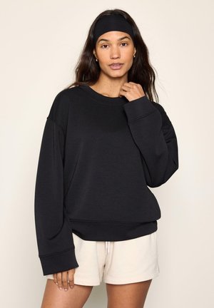 Frau mit langen dunklen Haaren, die ein schwarzes Stirnband, einen schwarzen oversized Sweatshirt und hellbeige Shorts trägt, steht vor einem einfarbigen Hintergrund.