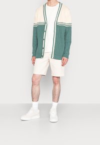 Cardigan de canalé en color verde azulado y crema con escote en V, cierre de botones y acentos a rayas, combinado con pantalones cortos color crema y zapatillas blancas.