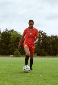 Oranžový futbalový dres s bielymi akcentmi, s emblémom leva, čiernymi ponožkami a viacfarebným futbalovým loptou na trávnatej ploche.