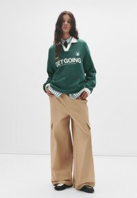 Zelený rebrovaný sveter s bielym textom, vrstvený cez pruhovanú košeľu s golierom. Široké béžové cargo nohavice s bočnými vreckami a čiernymi topánkami.