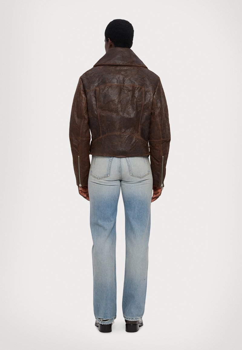 Brown leather biker jacket with a cropped cut and wide collar, featuring detailed stitching and silver zippered accents. Worn with light blue jeans.
