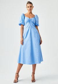 Hele sinine midi kleit puff-sleeved, rikka keha ja voolava A-löökskurtiga. Valmistatud siledast, kergest kangast. Mustad rihmadega kõrged kontsad.