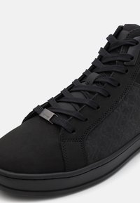 Sorte høye sneakers med et strukturert mønster, som har en glatt overflate, sorte lisser og en metalllogo-detalj på siden.
