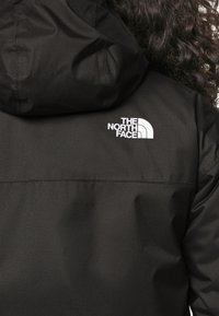 Zwarte capuchonjack van textuurstof met een wit logo van "The North Face" op de bovenrug.