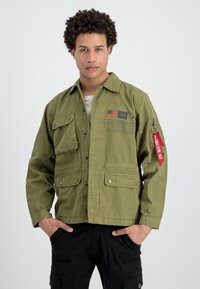 Chaqueta ligera de color verde oliva con múltiples bolsillos frontales, cierres a presión y una etiqueta roja en la manga. Presenta gráficos e inscripciones impresas.