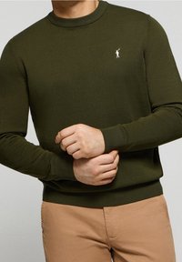 Olivgrön stickad tröja med rundad halsringning, ribbade ärmslut och nederkant, med en liten vit logotyp på bröstet.