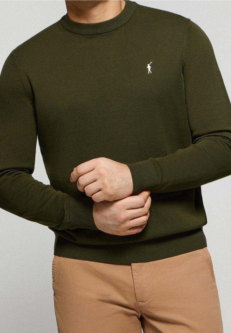 Olivgrön stickad tröja med rundad halsringning, ribbade ärmslut och nederkant, med en liten vit logotyp på bröstet.