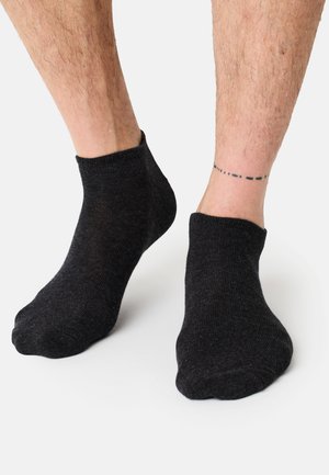 Schwarze Knöchelsocken aus einem weichen, strukturierten Stoff mit geripptem Bündchen und enger Passform, getragen an nackten Füßen mit sichtbarem Beinhaar und einem Tattoo.
