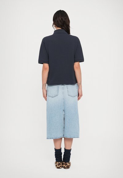 Γυναίκα με σκούρα μαλλιά που φοράει σκούρο κοντομάνικο πόλο μπλουζάκι, ανοιχτό μπλε τζιν μεσαίου μήκους φούστα, σκούρες κάλτσες και παντόφλες με λεοπάρ μοτίβο, όρθια γυρισμένη με την πλάτη προς το μέρος μας.