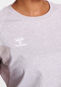 Jasnoszara bluza z okrągłym dekoltem i rękawami raglanowymi. Posiada gładką fakturę oraz białe logo Hummel nadrukowane z przodu.