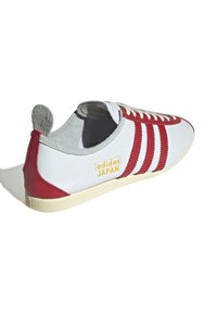 Vit sneaker med röda accenter, tre ränder och en gul "adidas JAPAN"-logga. Har en gummisula och en texturerad grå tunga.