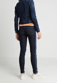 Person wearing dark blue denim jacket and skinny jeans with white sneakers, standing against a plain light background.