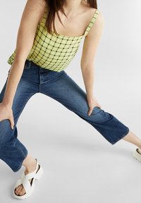 Limegrünes Karomuster-Trägershirt mit dünnen Trägern, kombiniert mit verkürzten blauen Jeans und weißen Sandalen mit dicker Sohle.