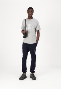Tricou din bumbac gri cu imprimeu alb și pantaloni de trening bleumarin. Geantă de umăr neagră și adidași închiși cu detalii texturate. Design simplu.