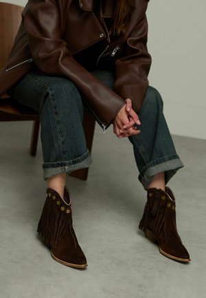 Bruine suede enkellaarzen hebben franjes en decoratieve gouden knopen. De laarzen hebben een spitse neus en een lage, gestapelde hak.