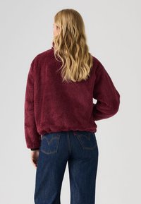 Bordo flis jakna z mehko, teksturirano tkanino, ohlapnega kroja z elastičnim pasom, nošena preko temnih jeansa.