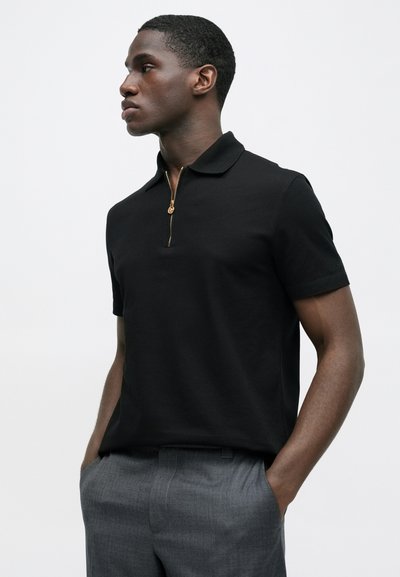 Juoda polo marškinėliai trumpomis rankovėmis, su auksiniu užtrauktuku ir mažu siuvinėtu detalė ant rankovės, pagaminti iš lygios medžiagos.