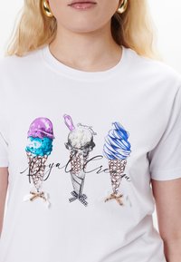Hvid t-shirt med tre isvafler: lilla og blå kugler, grå med perle detaljer, og blå snoet. Inkluderer dekorative sløjfer.