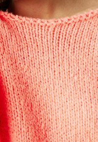 Dziewiński sweter w jasnym koralowym kolorze, z teksturowanym ryżowym wzorem. Dekolt jest szeroki z obszywanym brzegiem, wykonany z miękkiej, puszystej przędzy.